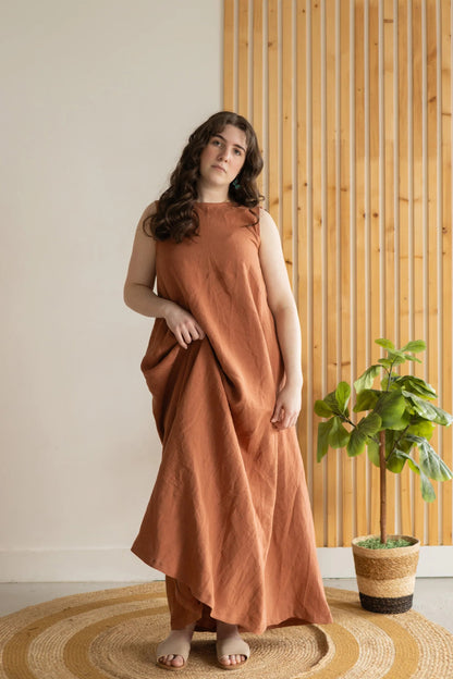A woman wearing the Caroline Dress by Lights of All in Sedona, a sleeveless dress linen maxi dress with a V-neck at the back. She is standing against a white and wood panelled wall. 
