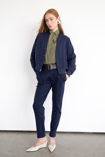 A woman wearing the Ponte di Roma Larimar Pants by Eve Lavoie in Navy, slim-fitting pull-on pants with a wide waistband, front pleats, and an ankle-length hem with a side slit detail. She is wearing them with a green shirt and navy jacket and standing against a white background. 