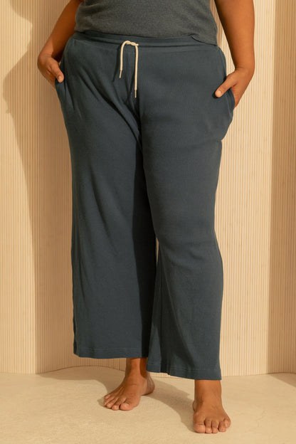Waist down view of a woman wearing the Organic Cotton Lounge Pants from Lights of All in Deep Sea, waffle knit pants with an elastic and drawstring waist, straight legs, and pockets. She is wearing them with a charcoal tank top and standing against a cream wall. 
