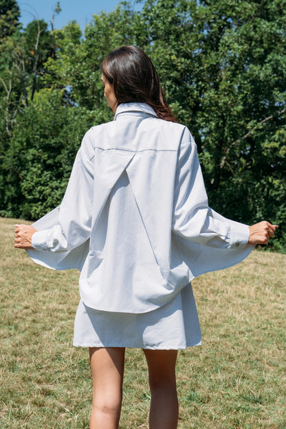 Back view of a woman wearing the Kyoto Blouse by MAS in Stripe, a classic collared button down with a cross-pleat at the back. She is wearing it open over a dress and standing outdoors. 