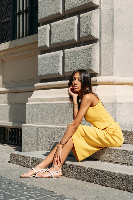 A woman in wearing the Lima Tank by MAS in Lemon, a cropped rib tank with a high neck and wide straps. She is wearing it with a matching skirt and sitting on stone steps outdoors.