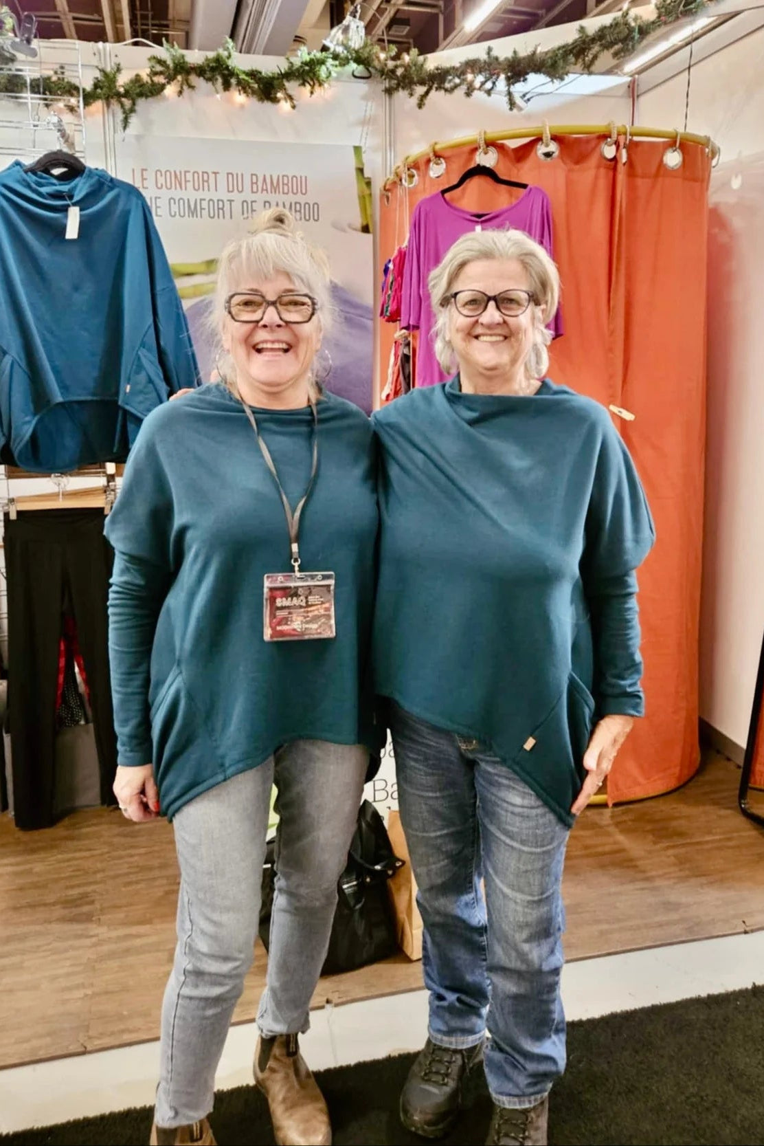 Two women wearing the Meeso Sweater by Moovment in Teal. The top has big side pockets, a wide mock turtleneck, and long dropped sleeves. They are wearing it with jeans and standing inside a clothing store.