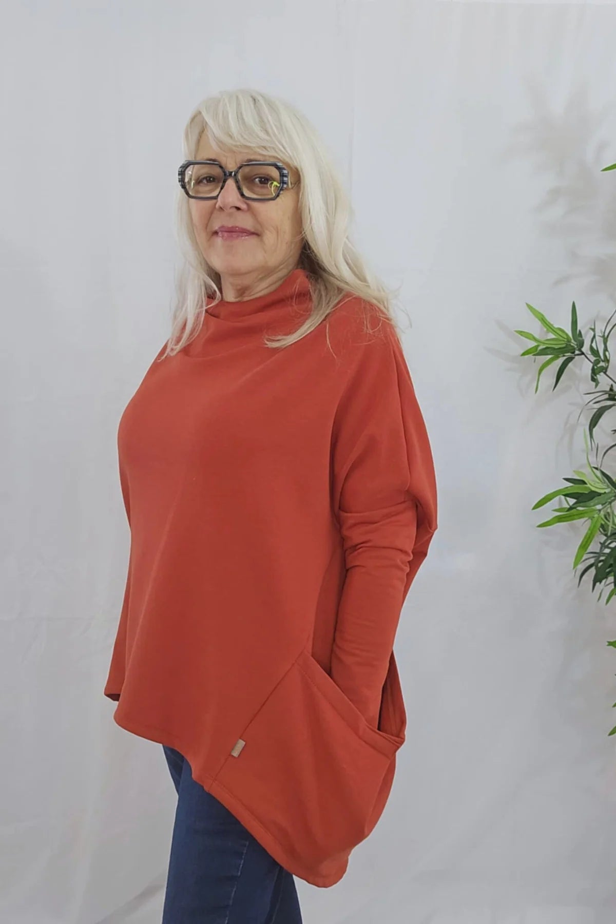 A woman wearing the Meeso Sweater by Moovment in Clay. The top has big side pockets, a wide mock turtleneck, and long dropped sleeves. She is wearing it with jeans and standing against a white wall with a plant beside her. 