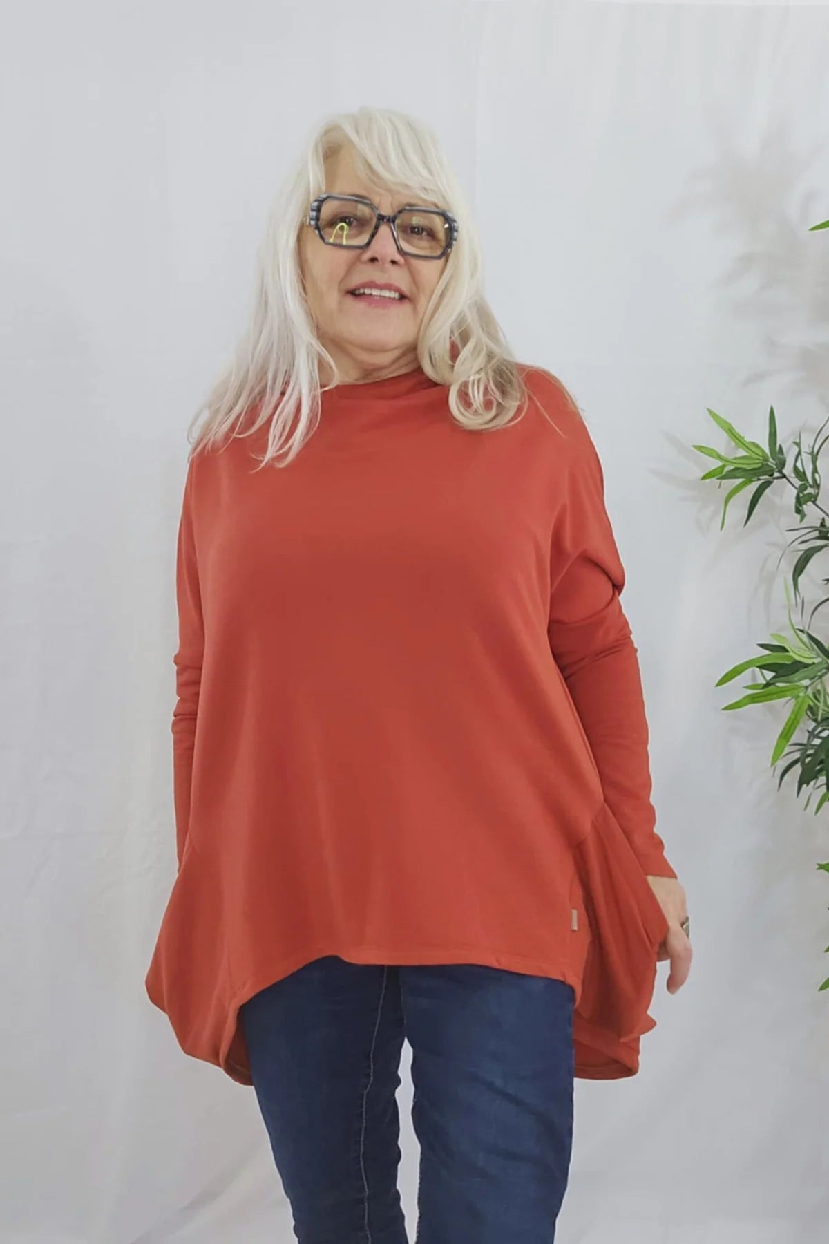A woman wearing the Meeso Sweater by Moovment in Clay. The top has big side pockets, a wide mock turtleneck, and long dropped sleeves. She is wearing it with jeans and standing against a white wall with a plant beside her. 