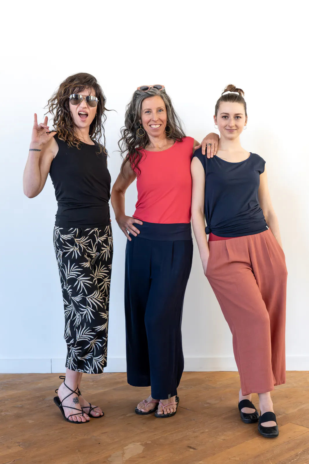 Three women wearing the Sol Top from Marie C, a reversible cap sleeve tee. From left to right they are wearing it in Black, Coral and Navy. They are all wearing pants and are standing in a white room with a wooden floor.