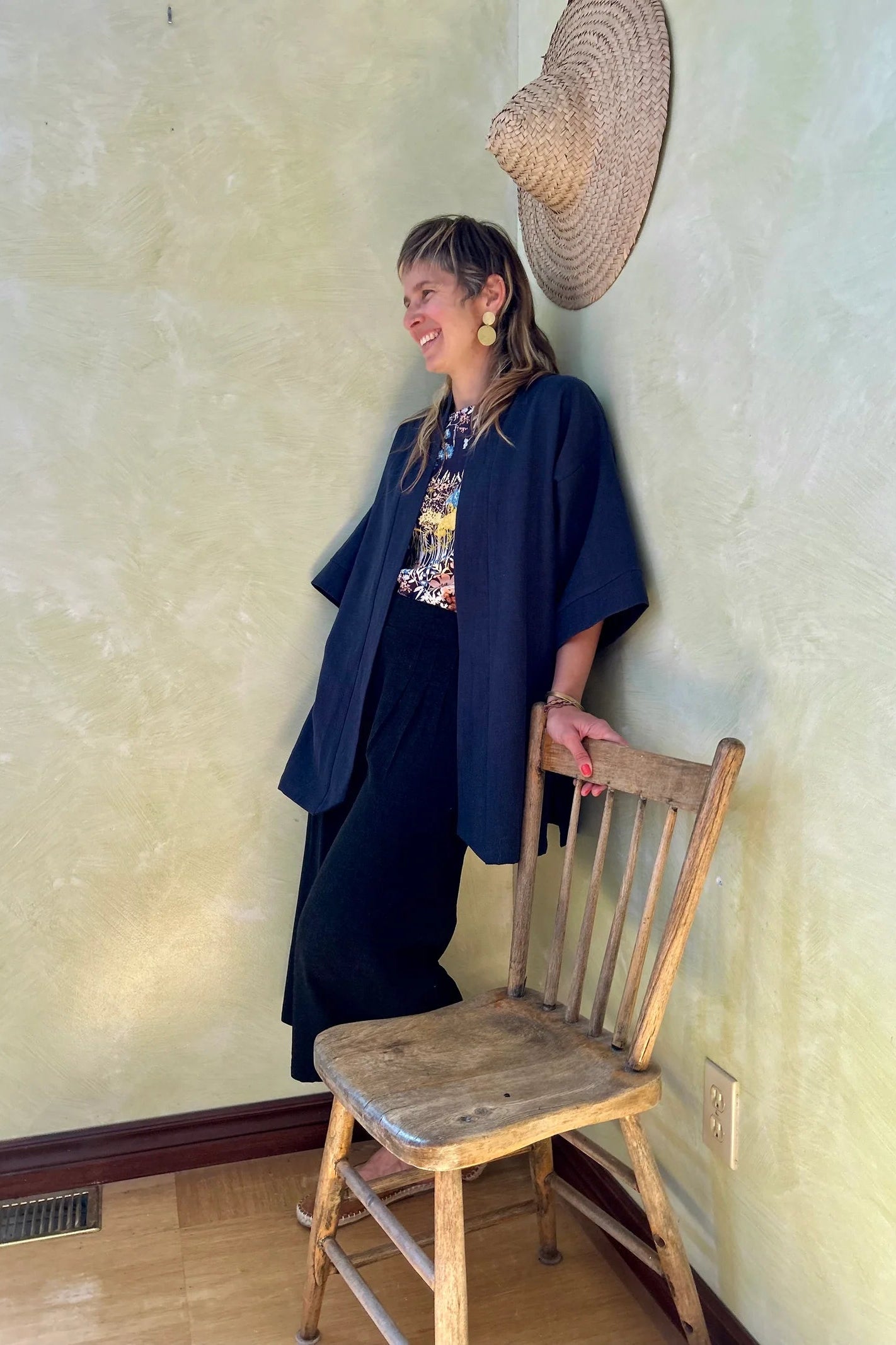 A woman wearing the Mirano Jacket by Kazak in Indigo, a long kimono style jacket with wide 3/4 sleeves. She is wearing it with the Boronia top in Prairie print and navy pants and standing next to a wooden chair against a textured wall.