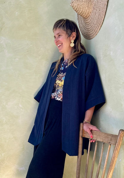A woman wearing the Mirano Jacket by Kazak in Indigo, a long kimono style jacket with half sleeves. She is wearing it with the Boronia top in Prairie print and navy pants and standing next to a wooden chair against a textured wall.