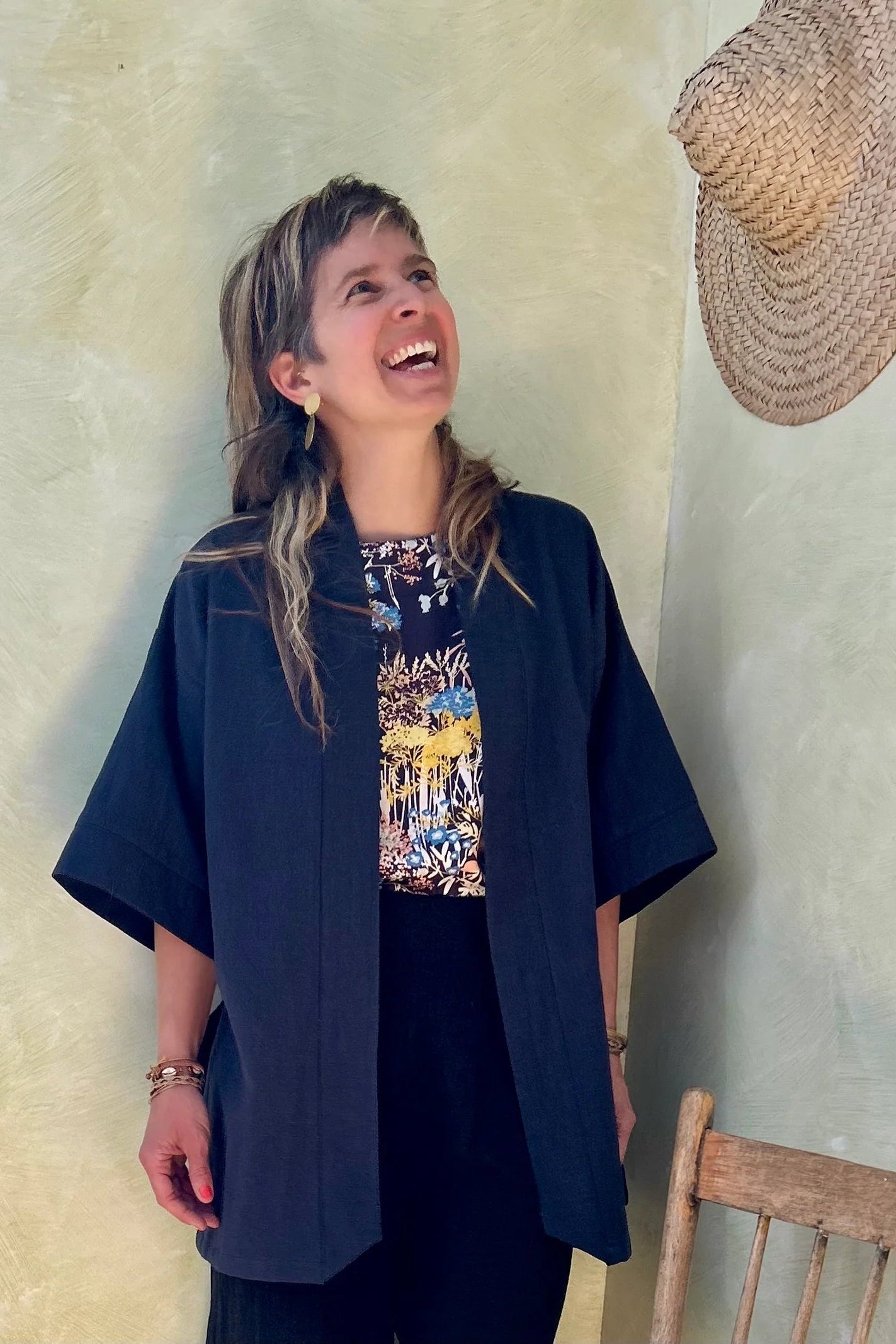 A woman wearing the Mirano Jacket by Kazak in Indigo, a long kimono style jacket with wide 3/4 sleeves. She is wearing it with the Boronia top in Prairie print and navy pants and standing next to a wooden chair against a textured wall.