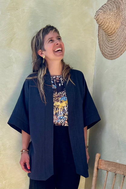 A woman wearing the Mirano Jacket by Kazak in Indigo, a long kimono style jacket with wide 3/4 sleeves. She is wearing it with the Boronia top in Prairie print and navy pants and standing next to a wooden chair against a textured wall.