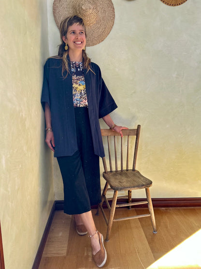 A woman wearing the Mirano Jacket by Kazak in Indigo, a long kimono style jacket with wide 3/4 sleeves. She is wearing it with the Boronia top in Prairie print and navy pants and standing next to a wooden chair against a textured wall.