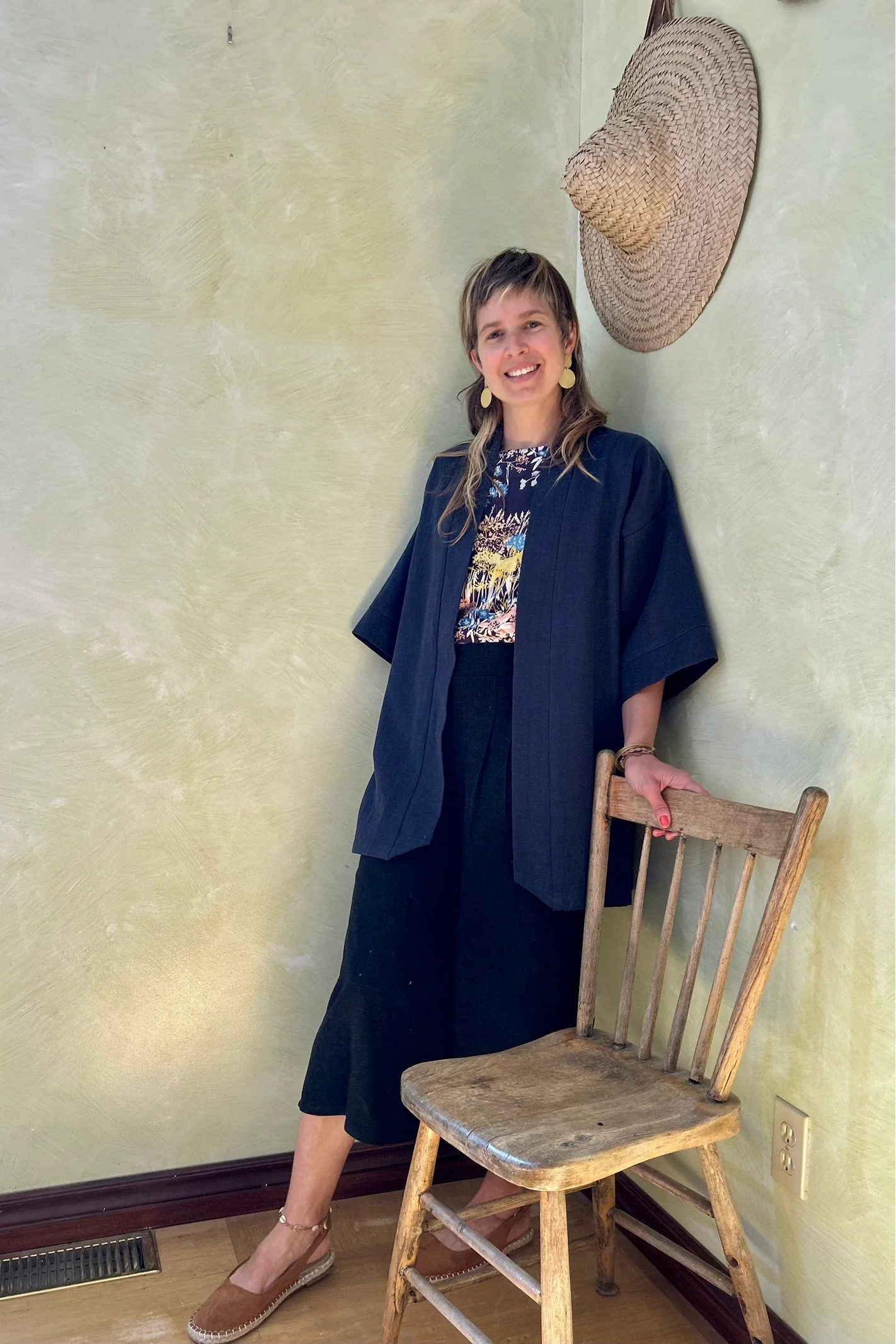A woman wearing the Mirano Jacket by Kazak in Indigo, a long kimono style jacket with half sleeves. She is wearing it with the Boronia top in Prairie print and navy pants and standing next to a wooden chair against a textured wall.
