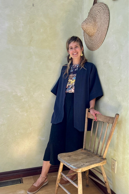 A woman wearing the Mirano Jacket by Kazak in Indigo, a long kimono style jacket with half sleeves. She is wearing it with the Boronia top in Prairie print and navy pants and standing next to a wooden chair against a textured wall.