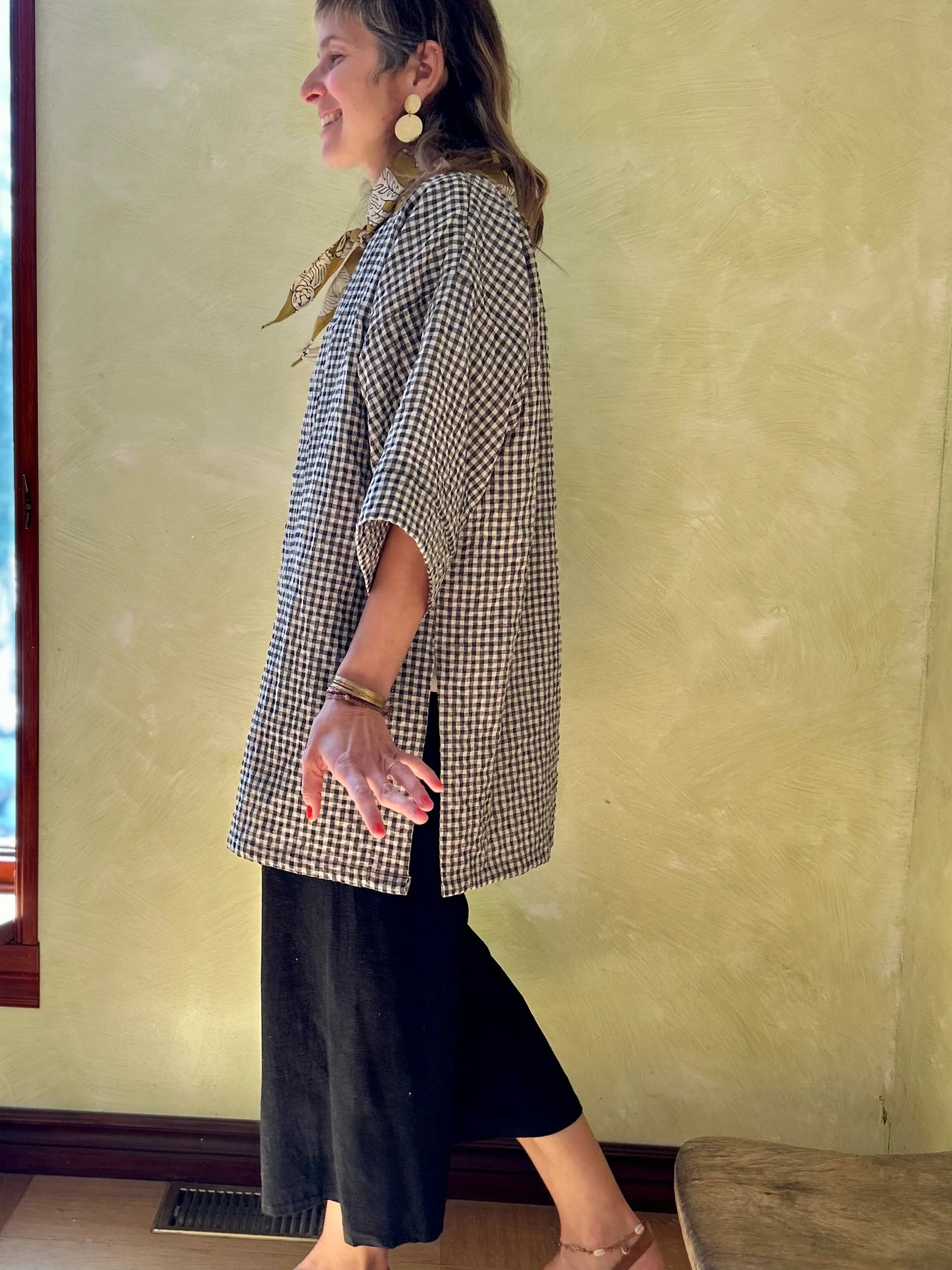 Side view of a woman wearing the Mirano Jacket by Kazak in Teal Vichy Teal, a long kimono style jacket with half sleeves. She is wearing it with the a white top and navy pants and standing next to a wooden chair against a textured wall.
