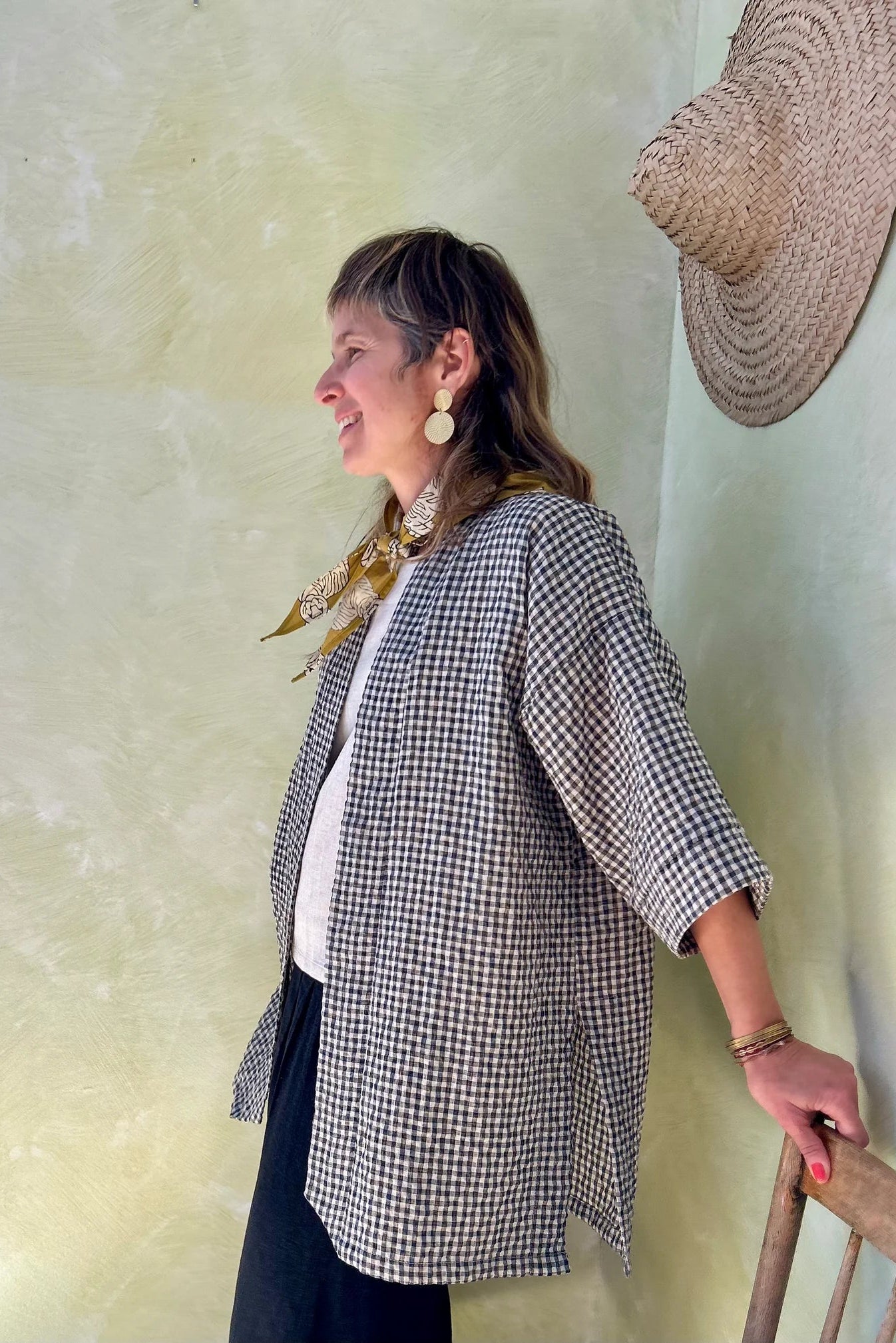 Side view of a woman wearing the Mirano Jacket by Kazak in Teal Vichy Teal, a long kimono style jacket with half sleeves. She is wearing it with the a white top and navy pants and standing next to a wooden chair against a textured wall.