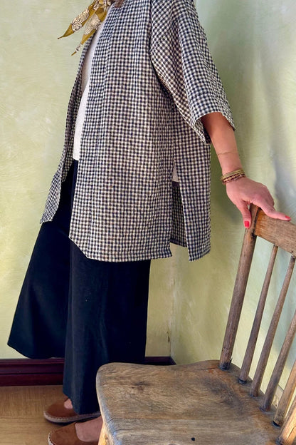 Neck down view of a woman wearing the Mirano Jacket by Kazak in Teal Vichy Teal, a long kimono style jacket with half sleeves. She is wearing it with the a white top and navy pants and standing next to a wooden chair against a textured wall.