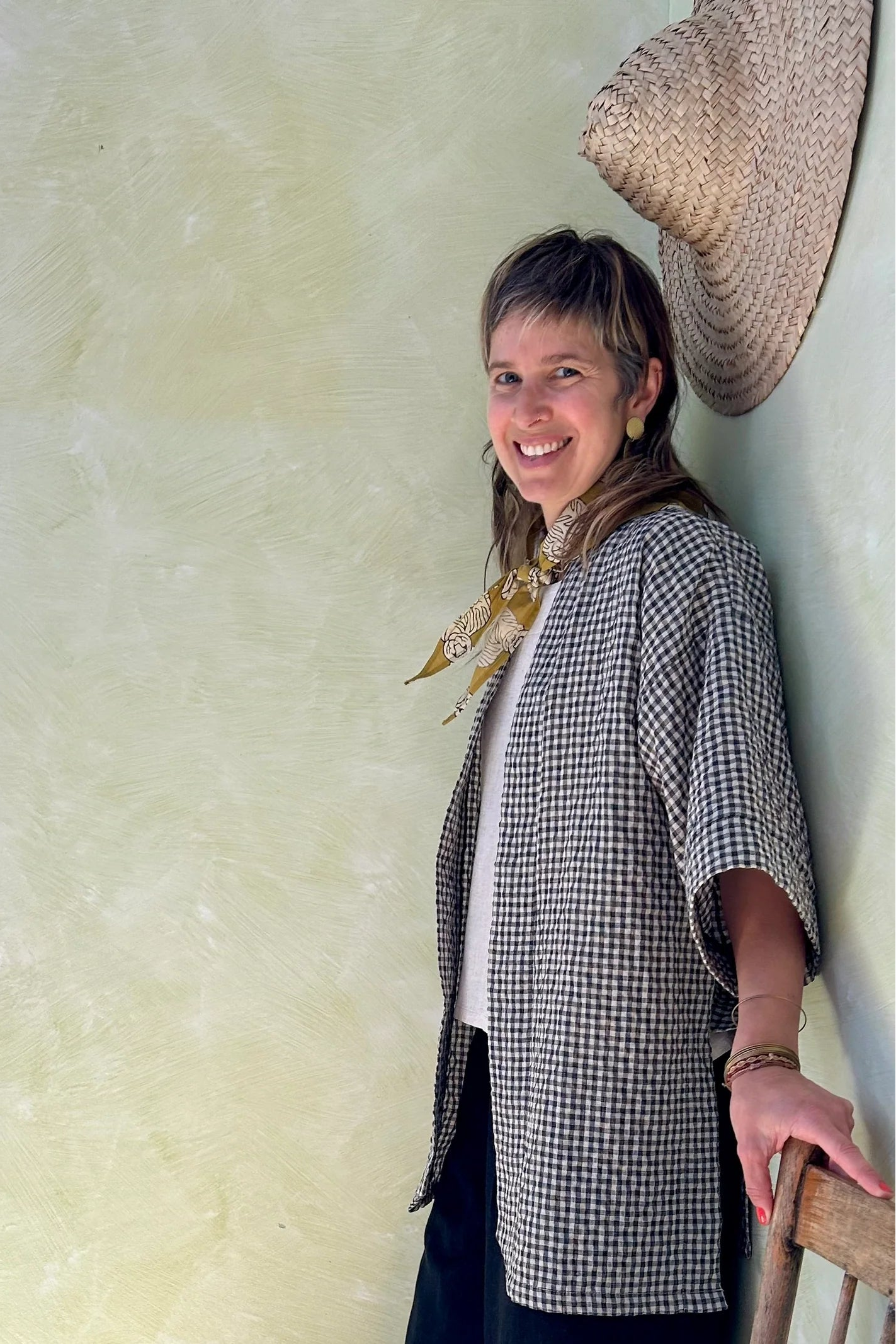 A woman wearing the Mirano Jacket by Kazak in Teal Vichy Teal, a long kimono style jacket with half sleeves. She is wearing it with the a white top and navy pants and standing next to a wooden chair against a textured wall.
