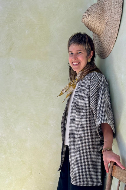 A woman wearing the Mirano Jacket by Kazak in Teal Vichy Teal, a long kimono style jacket with half sleeves. She is wearing it with the a white top and navy pants and standing next to a wooden chair against a textured wall.