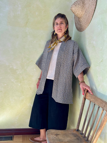 A woman wearing the Mirano Jacket by Kazak in Teal Vichy Teal, a long kimono style jacket with half sleeves. She is wearing it with the a white top and navy pants and standing next to a wooden chair against a textured wall.