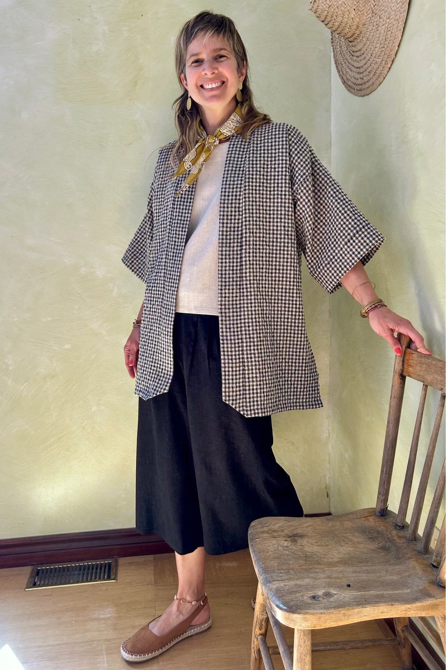 A woman wearing the Mirano Jacket by Kazak in Teal Vichy Teal, a long kimono style jacket with half sleeves. She is wearing it with the a white top and navy pants and standing next to a wooden chair against a textured wall.