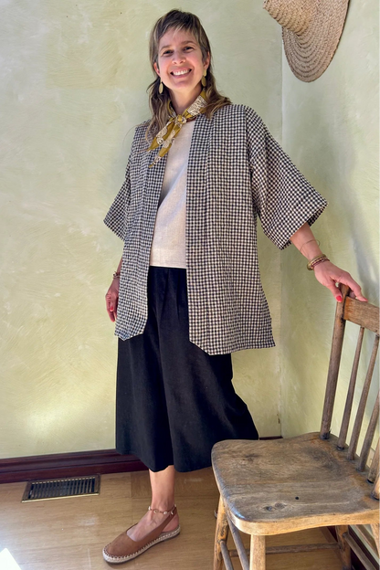A woman wearing the Mirano Jacket by Kazak in Teal Vichy Teal, a long kimono style jacket with half sleeves. She is wearing it with the a white top and navy pants and standing next to a wooden chair against a textured wall.