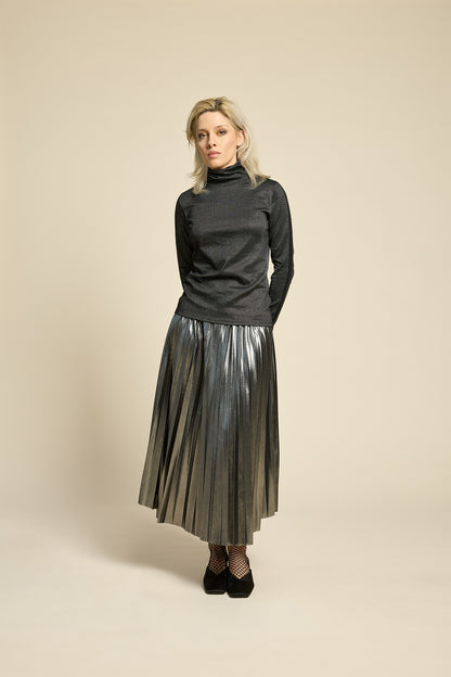 A woman wearing the Mortel Top by Cokluch in Facet Black, a metallic turtleneck with long sleeves. She is wearing it with the Feyer Skirt in Glow Knit Silver and standing in front of a beige background. 