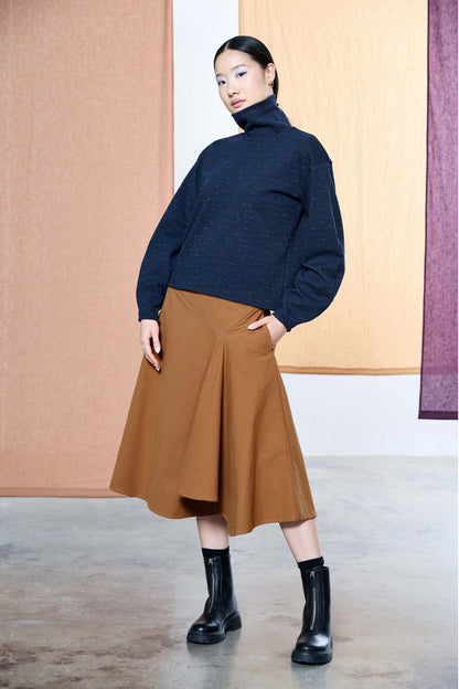 A woman wearing the Nathalie Top by Melow in Navy, a relaxed fitting turtleneck with long barrel sleeves and a speckled texture. She is wearing it with a copper skirt and standing in front of a pastel background. 