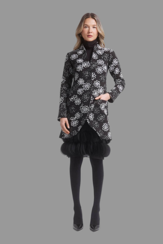 A woman wearing the Laurie Jacket by Dinh Ba in Black and White Floral, a thigh-length coat with V-neck, button front, patch pocket and curved front hem. She is wearing it with a black dress and standing in front of a grey background. 