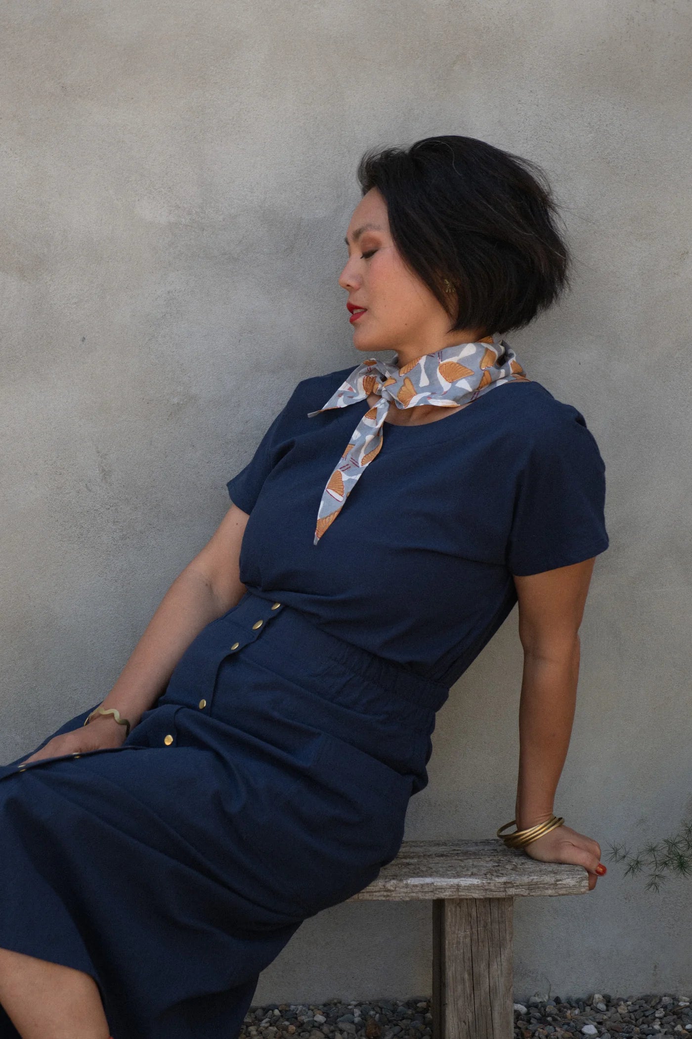 A woman wearing the Brendola Top from Kazak in Navy, a slightly cropped short sleeved top with three brass snaps that can be worn at the back or the front. She is wearing it with a scarf and the Nymphea skirt in Navy and sitting outside in front of a grey wall.