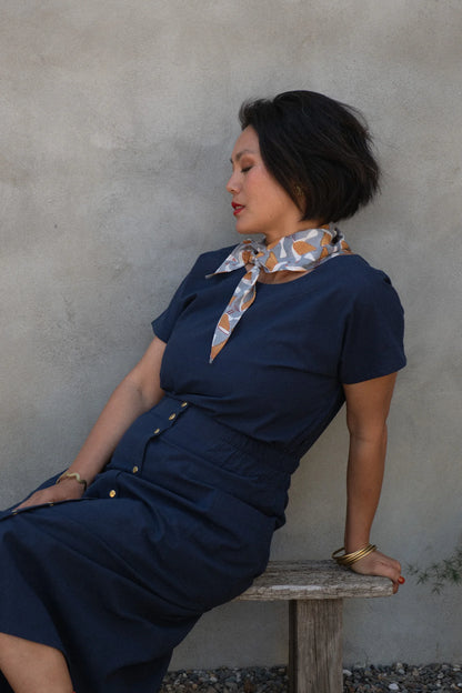 A woman wearing the Brendola Top from Kazak in Navy, a slightly cropped short sleeved top with three brass snaps that can be worn at the back or the front. She is wearing it with a scarf and the Nymphea skirt in Navy and sitting outside in front of a grey wall.