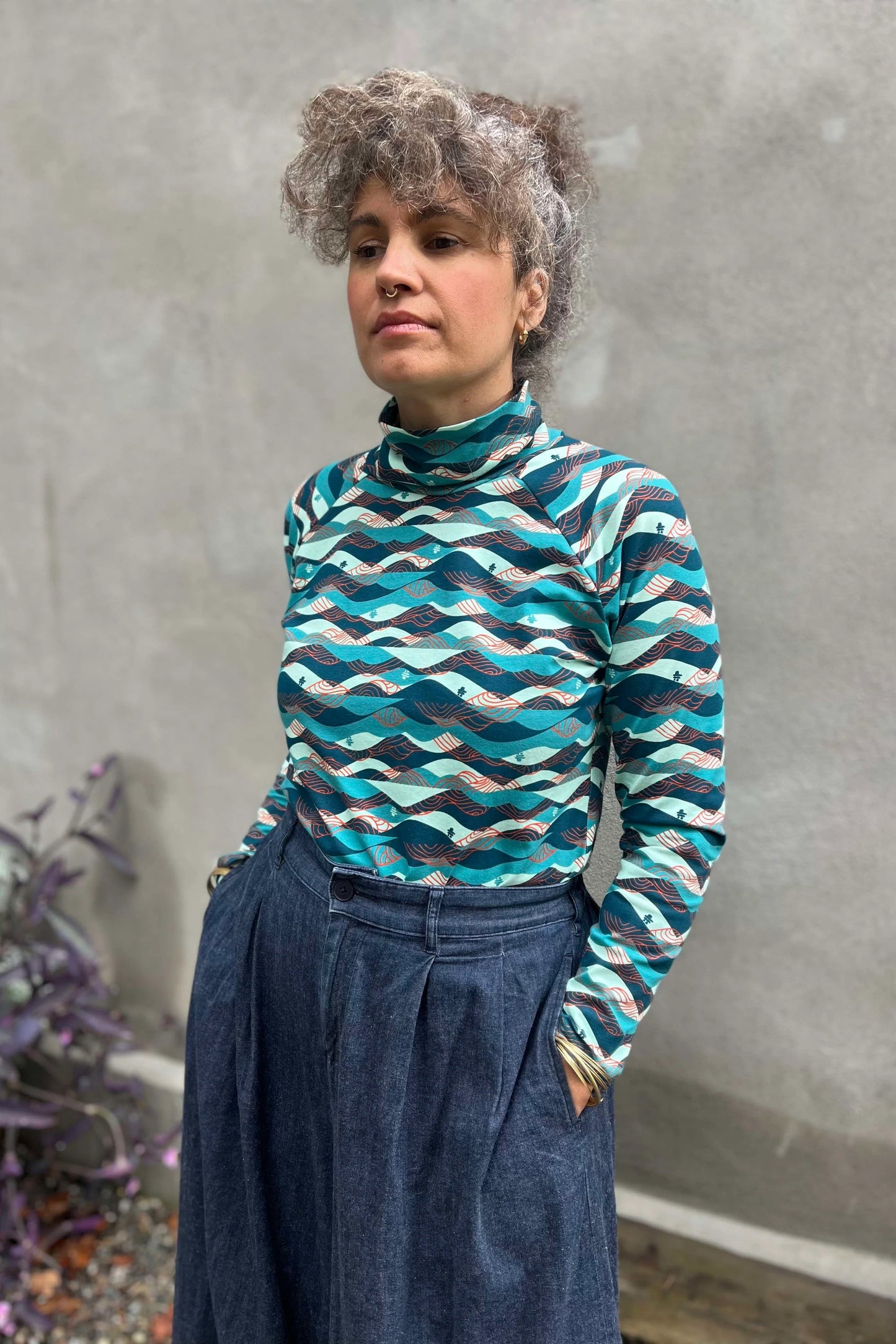 A woman wearing the Oris Raglan Turtleneck by Kazak in Inuksuk print, with a pattern of rolling waves and tiny inukshuks in shades of blue. She is wearing it with jeans and standing outside. 