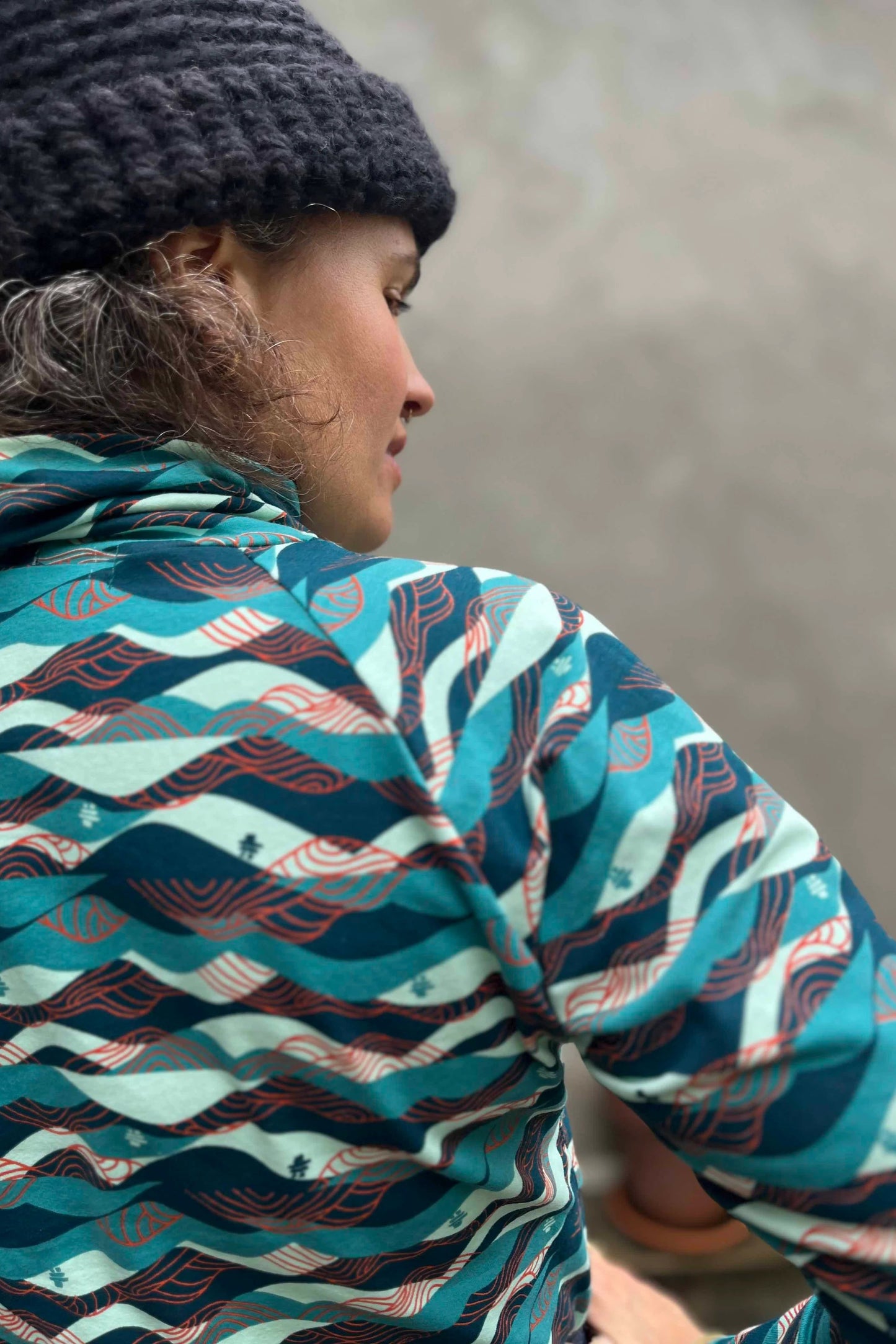 A woman wearing the Oris Raglan Turtleneck by Kazak in Inuksuk print, with a pattern of rolling waves and tiny inukshuks in shades of blue. 