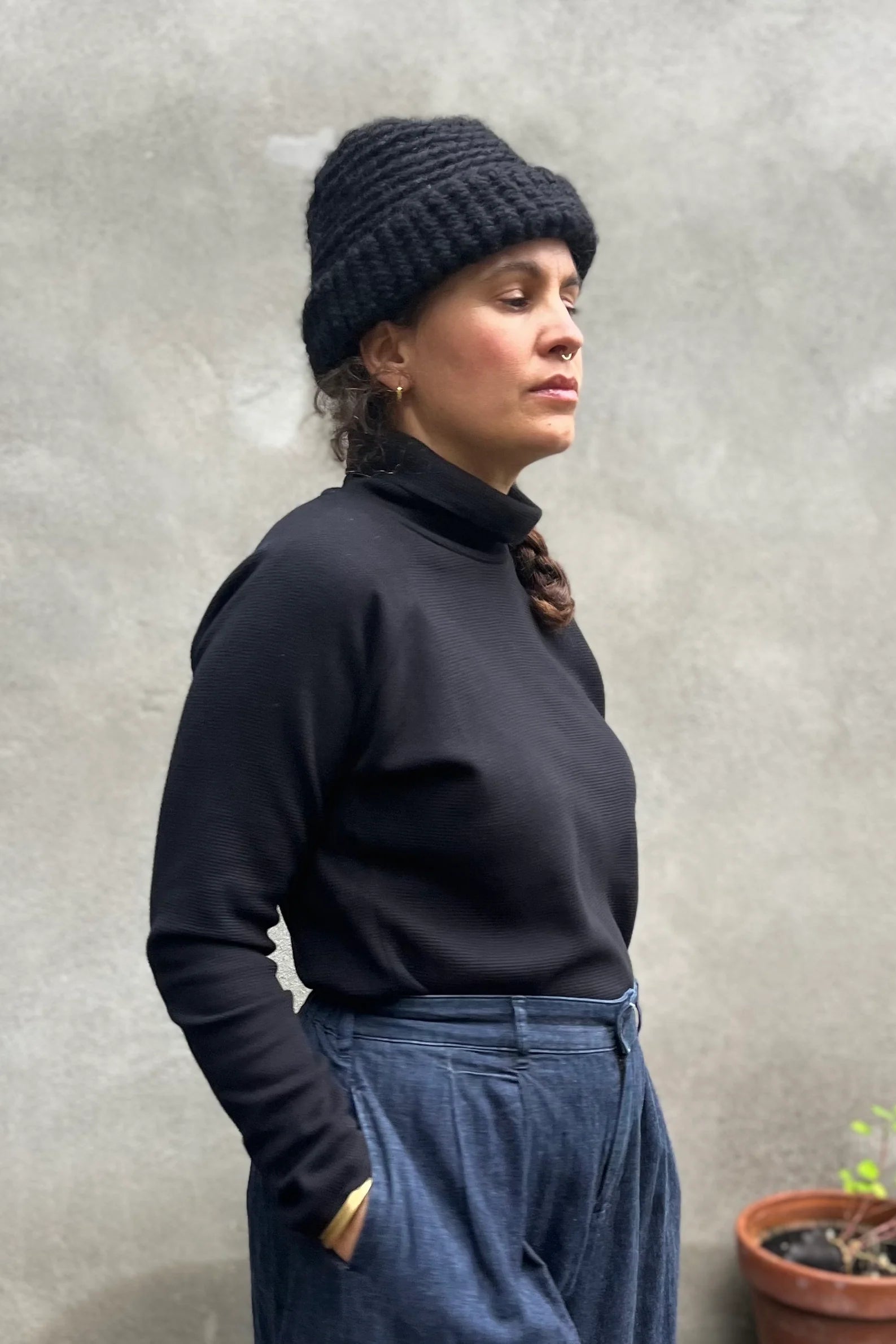 A woman wearing the Oris Raglan Turtleneck by Kazak in Black waffle. She is wearing it with jeans and standing outside. 