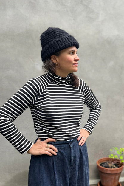 A woman wearing the Oris Raglan Turtleneck by Kazak in Black Stripe. She is wearing it with jeans and standing outside. 
