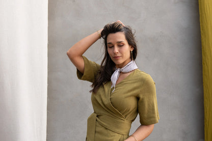 A woman wearing the Helleborus wrap Dress by Kazak in Mousse, standing outside by a white curtain and a cement wall. 