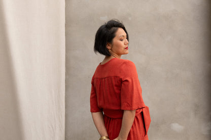 Back view of a woman wearing the Helleborus wrap Dress by Kazak in Paprika, standing outside by a white curtain and a cement wall. 
