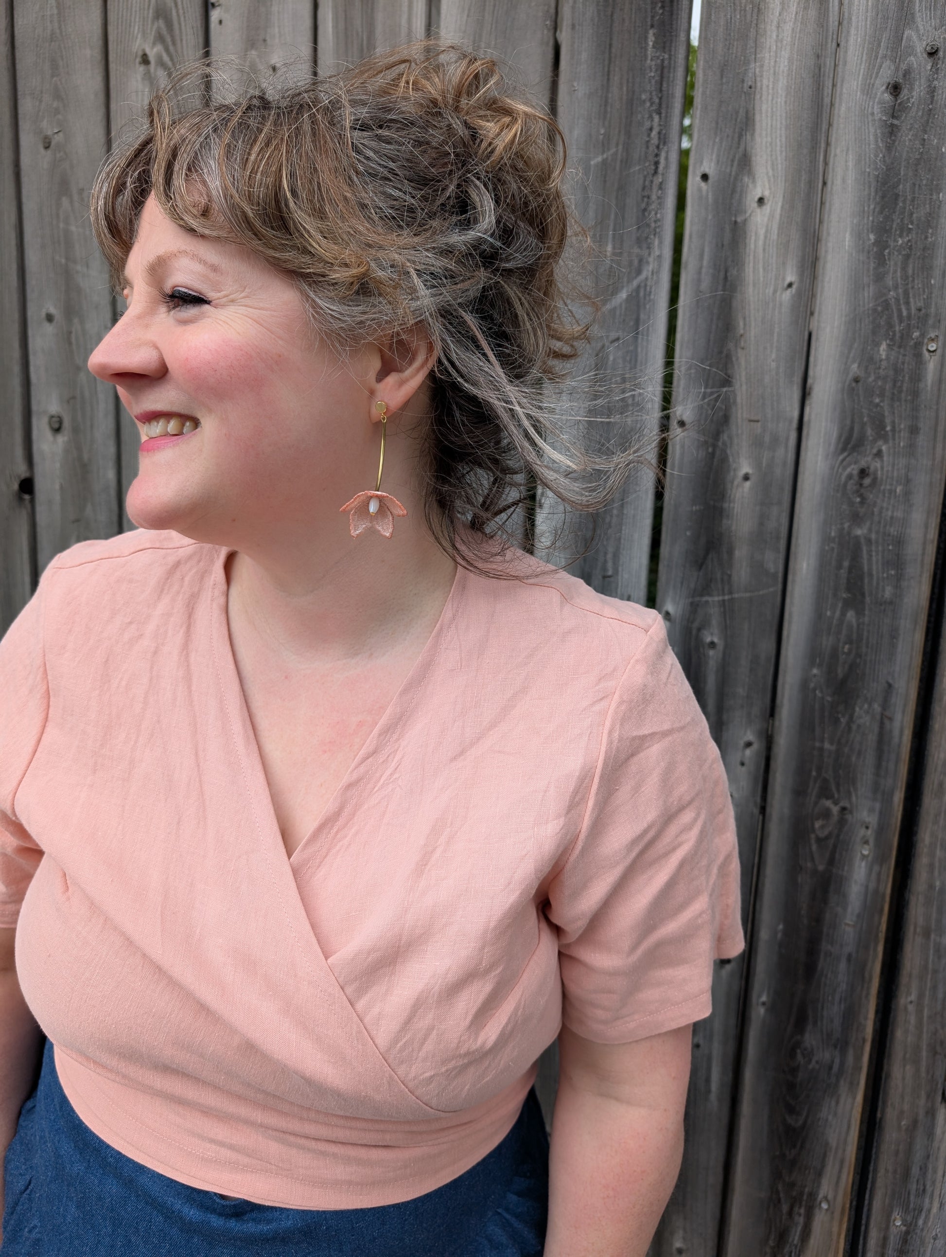 A woman wearing the Stella Top by Lights of All in Ballet Pink, a short sleeved linen wrap top that can be twisted or tied back or front. She is wearing it with the Margot Denim Skirt by Melow and standing by a fence. 