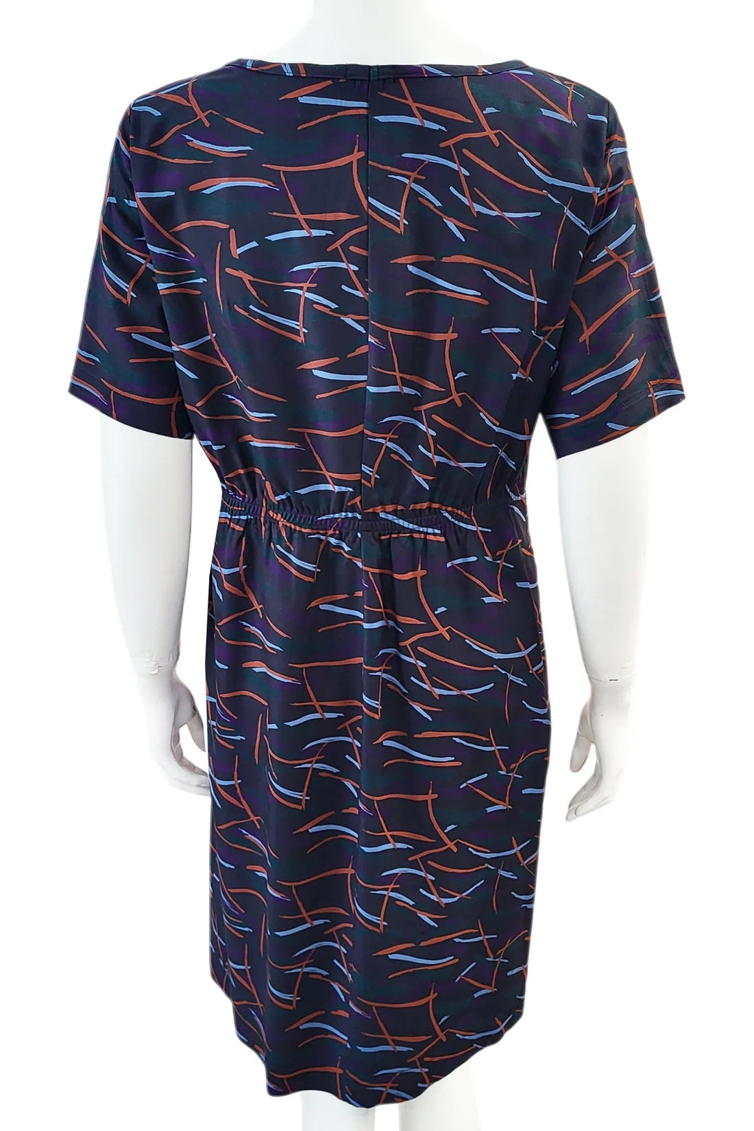 A back view of the Cecily Simple Shift Dress from Mandala in Dark Stix print is shown on a mannequin against a white background. It has a high neck, short sleeves, a knee-length hem, French darts, and elastic at the back of the waist. 