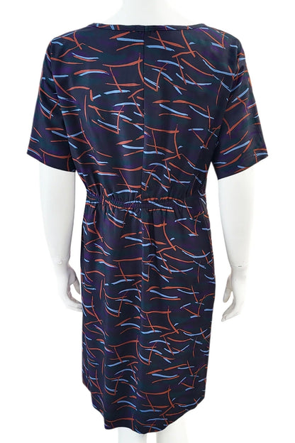 A back view of the Cecily Simple Shift Dress from Mandala in Dark Stix print is shown on a mannequin against a white background. It has a high neck, short sleeves, a knee-length hem, French darts, and elastic at the back of the waist. 