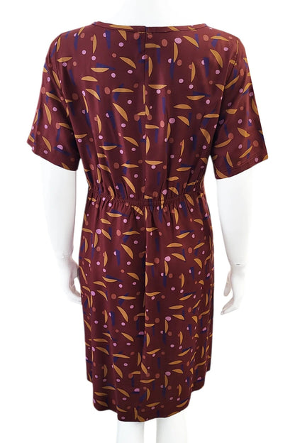 A back view of the Cecily Simple Shift Dress from Mandala in Playtime Rust print is shown on a mannequin against a white background. It has a high neck, short sleeves, a knee-length hem, French darts, and elastic at the back of the waist. 