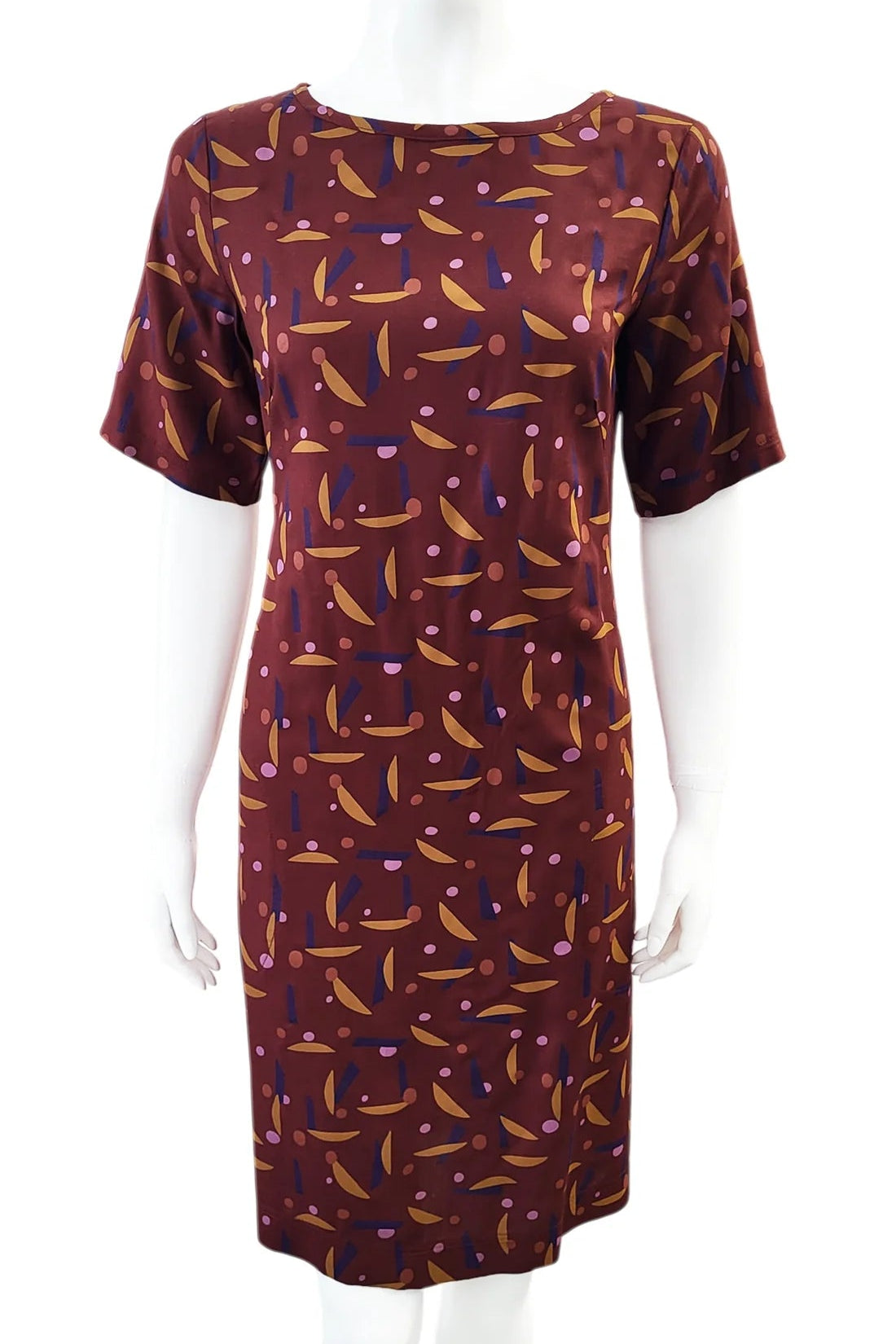 The Cecily Simple Shift Dress from Mandala in Playtime Rust print is shown on a mannequin against a white background. It has a high neck, short sleeves, a knee-length hem, French darts, and elastic at the back of the waist. 