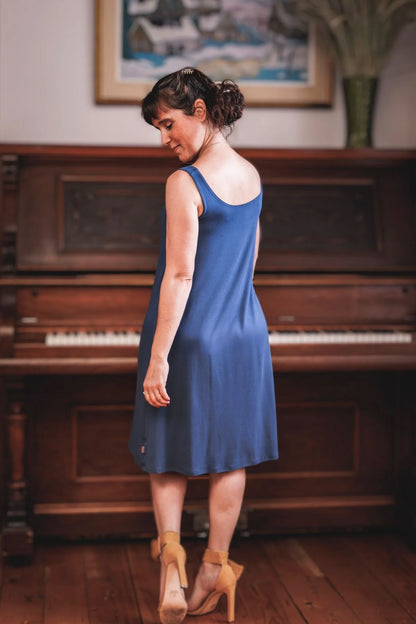 Back view of a woman wearing the Riana ami Dress by Moovment in Denim, a tank dress with a round neck front and back, A-line shape that flows away from the body, and knee-length hem, all in bamboo viscose. She is standing in front of a piano. 