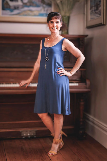 A woman wearing the Riana Cami Dress by Moovment in Denim, a tank dress with a round neck front and back, A-line shape that flows away from the body, and knee-length hem, all in bamboo viscose. She is standing in front of a piano. 