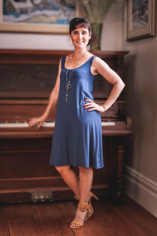 A woman wearing the Riana Cami Dress by Moovment in Denim, a tank dress with a round neck front and back, A-line shape that flows away from the body, and knee-length hem, all in bamboo viscose. She is standing in front of a piano. 