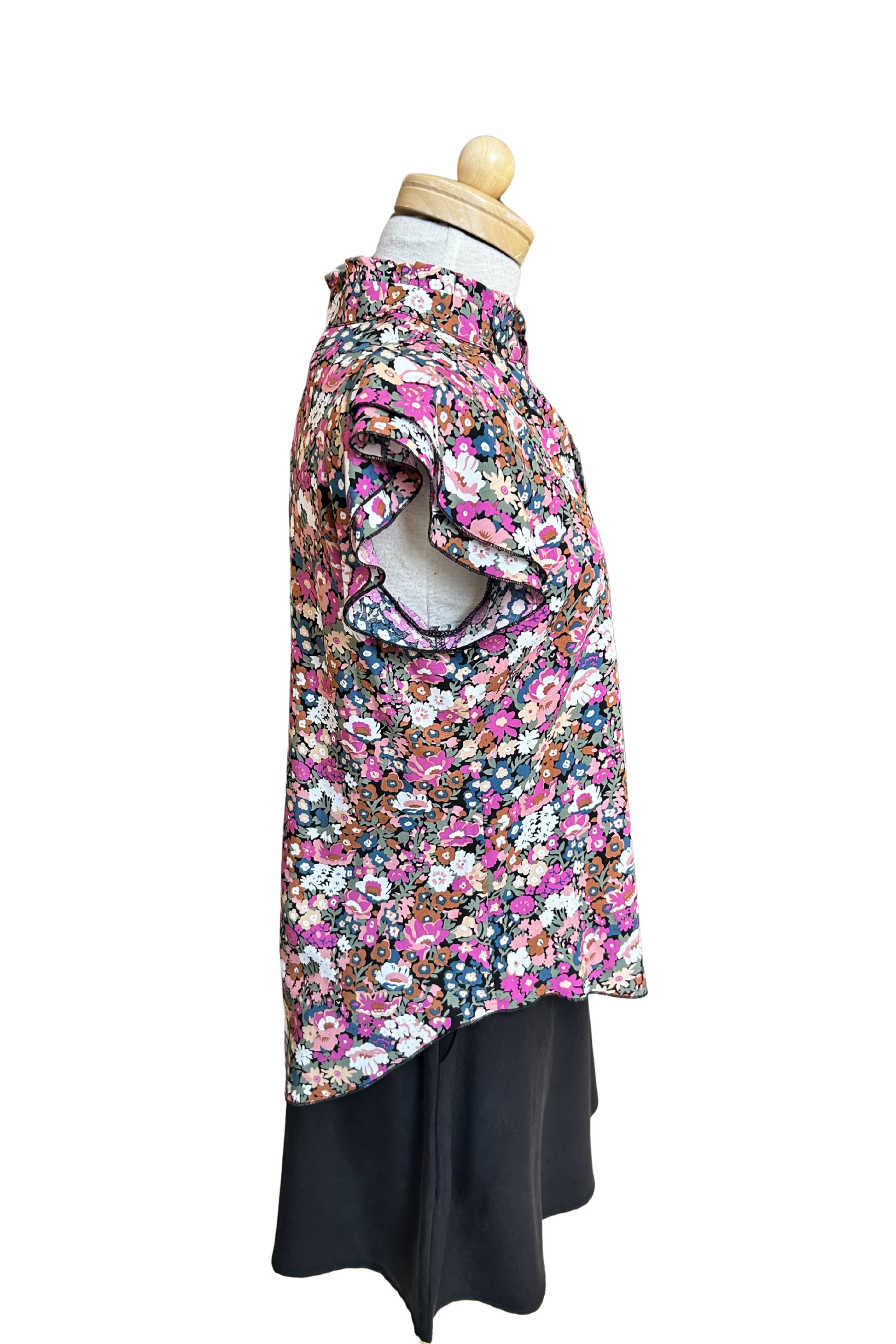 A side view of the Petal Blouse by Dotty in Pink/Blue floral print is shown on a mannequin against a white background. It has short floaty sleeves, a split V-neck, a ruffled collar, and a rounded hi-low hemline. 