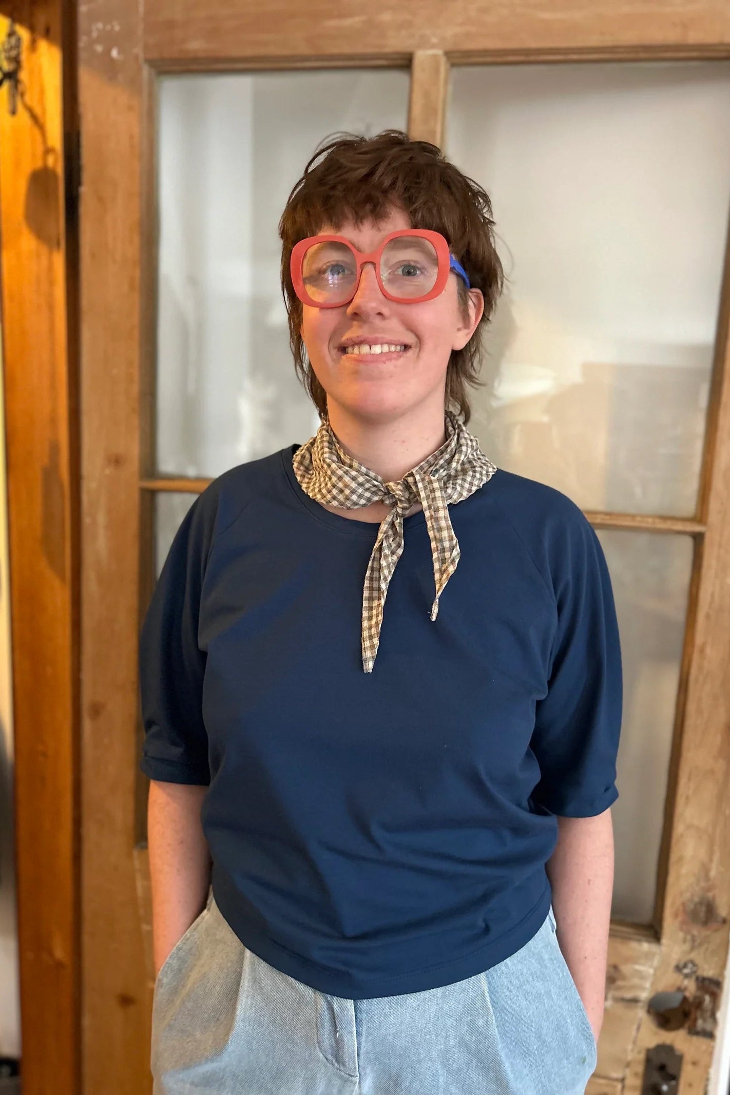 A woman wearing the Vernus Top by Kazak in Night with elbow-length raglan sleeves with a pleat detail at the shoulders, a round neck, and a fit that tapers at the waist. She is wearing it with light jeans and a checkered scarf and standing indoors by a glass-panelled door. 