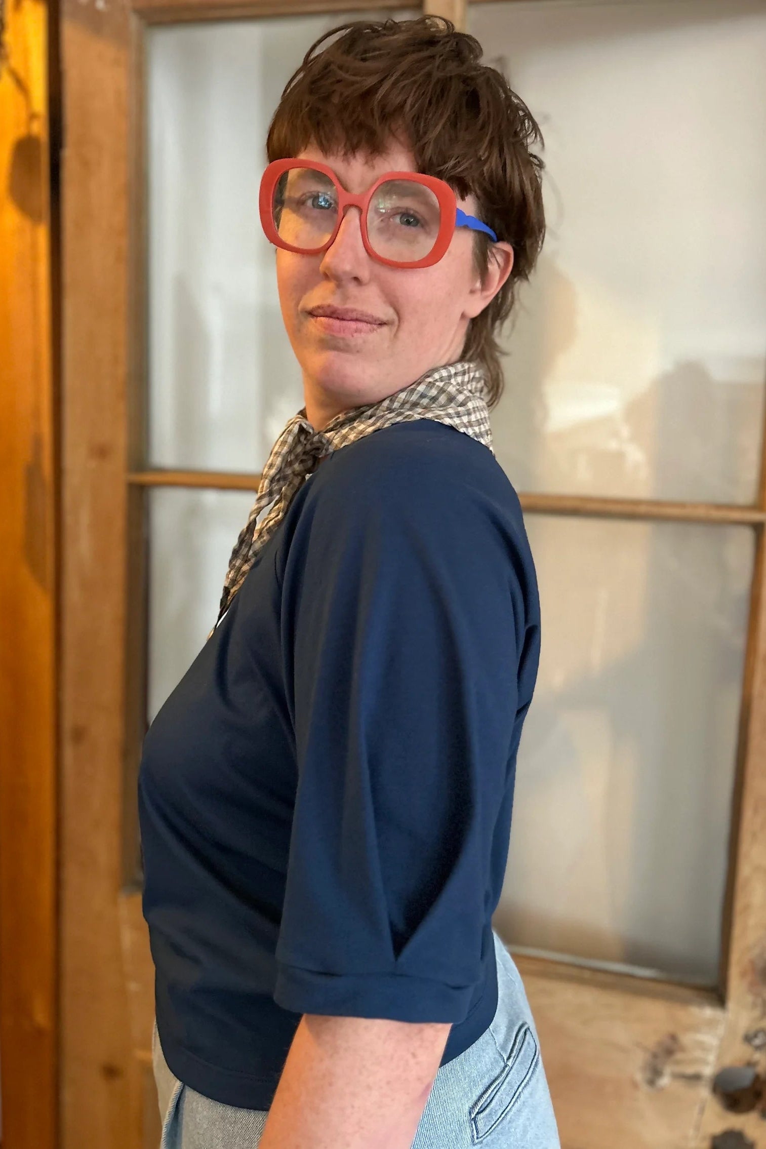 Side view of a woman wearing the Vernus Top by Kazak in Night with elbow-length raglan sleeves with a pleat detail at the shoulders, a round neck, and a fit that tapers at the waist. She is wearing it with light jeans and a checkered scarf and standing indoors by a glass-panelled door. 