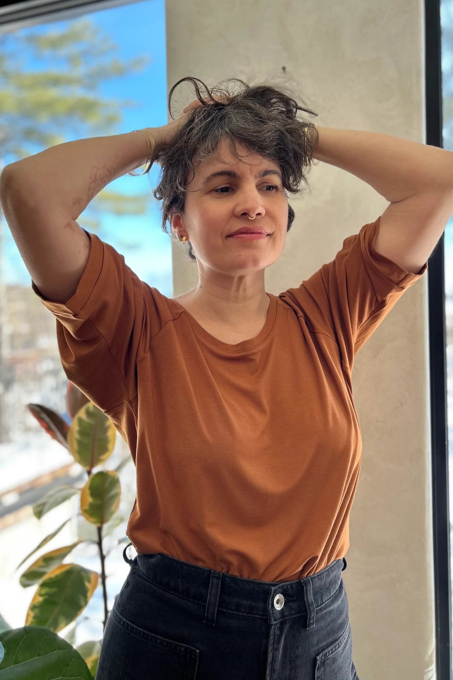 A woman wearing the Vernus Top by Kazak in Nutmeg, with elbow-length raglan sleeves with a pleat detail at teh shoulders, a round neck, and a fit that tapers at the waist. She is wearing it with dark pants indoors with a window in the background