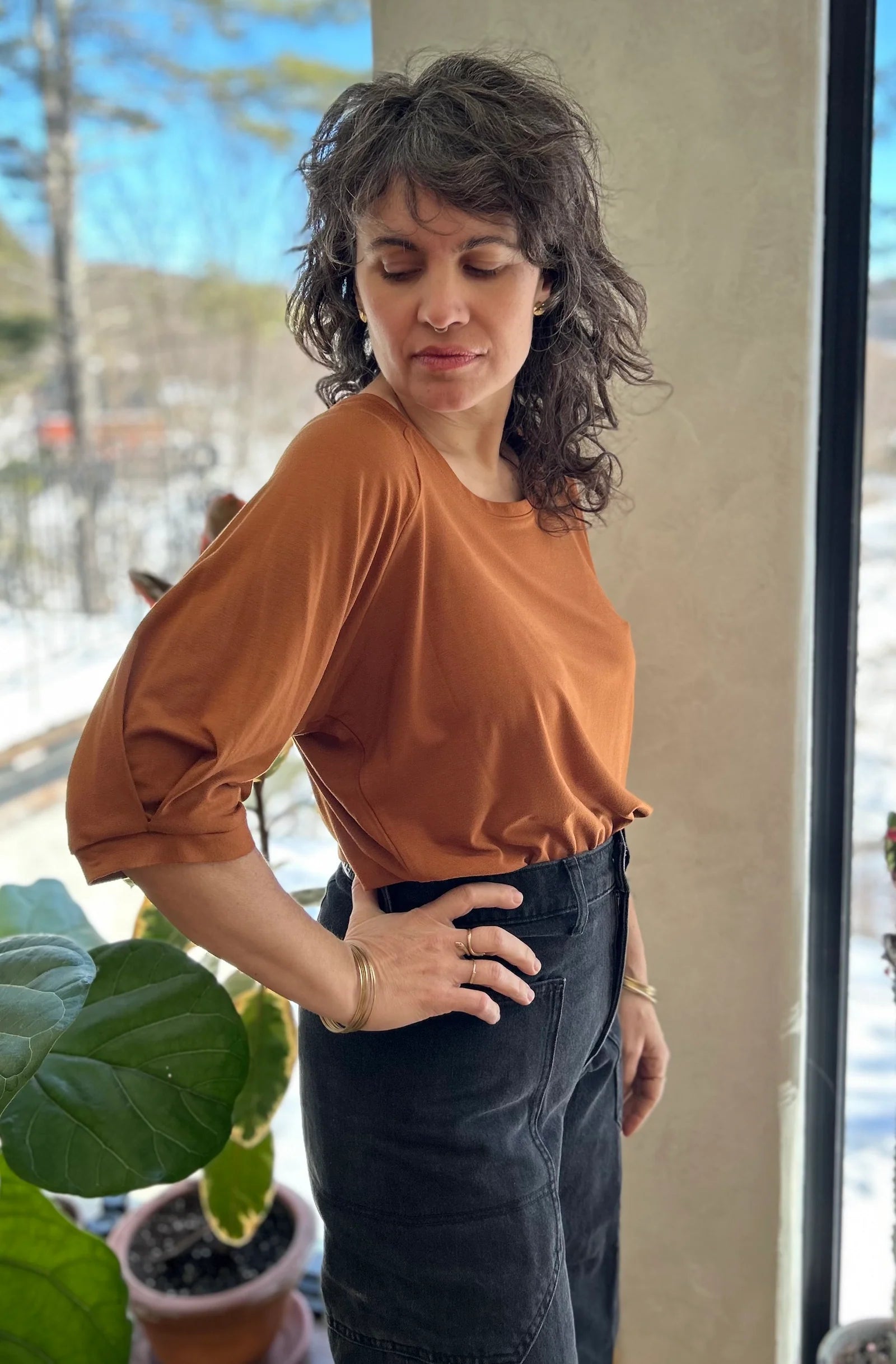 A woman wearing the Vernus Top by Kazak in Nutmeg, with elbow-length raglan sleeves with a pleat detail at teh shoulders, a round neck, and a fit that tapers at the waist. She is wearing it with dark pants indoors with a window in the background