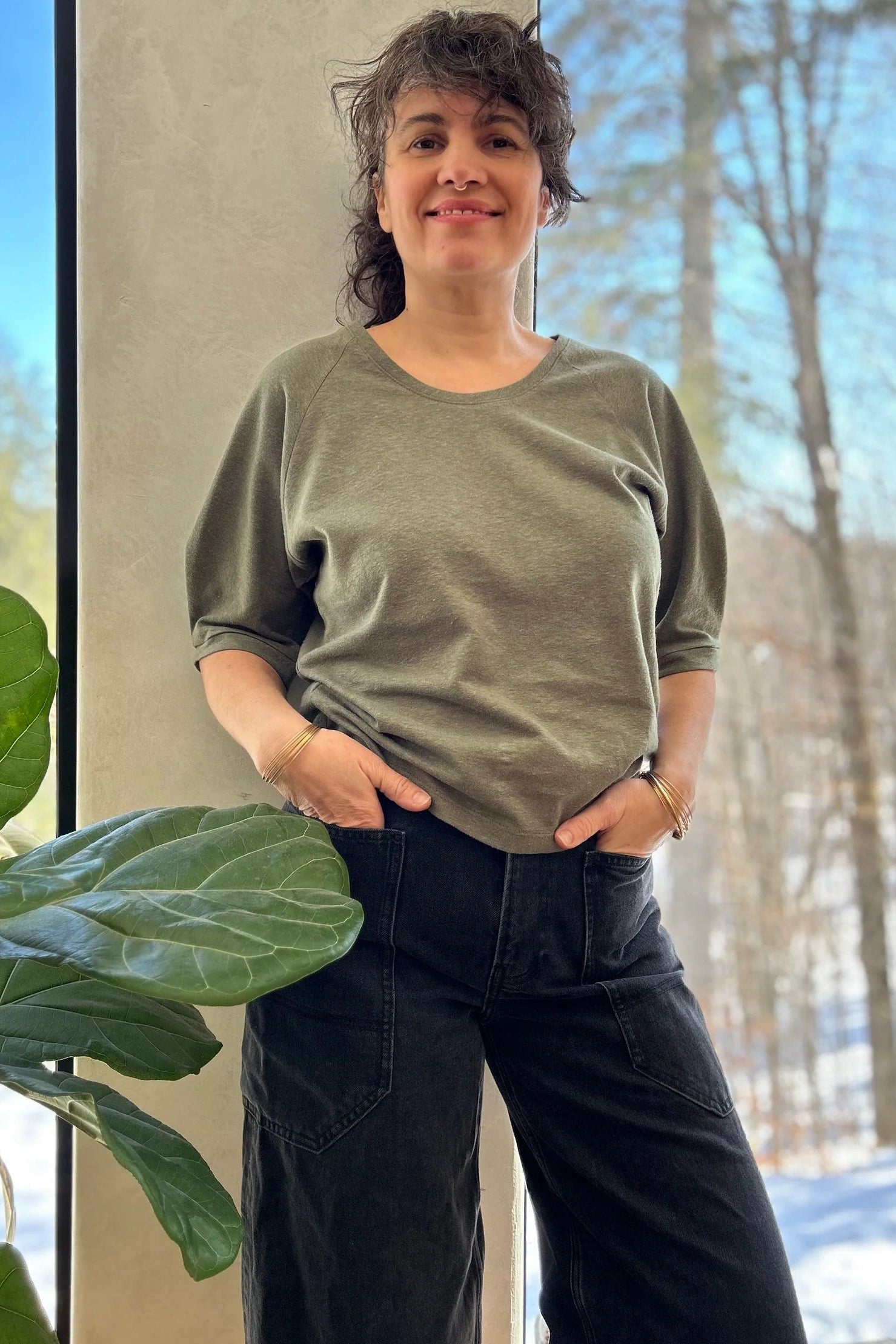A woman wearing the Vernus Top by Kazak in Sage with elbow-length raglan sleeves with a pleat detail at the shoulders, a round neck, and a fit that tapers at the waist. She is wearing it with dark jeans standing indoors by a window.