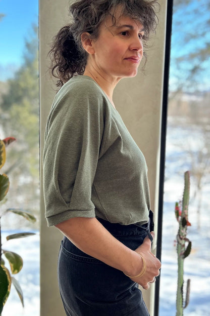 Side view of a woman wearing the Vernus Top by Kazak in Sage with elbow-length raglan sleeves with a pleat detail at the shoulders, a round neck, and a fit that tapers at the waist. She is wearing it with dark jeans standing indoors by a window.
